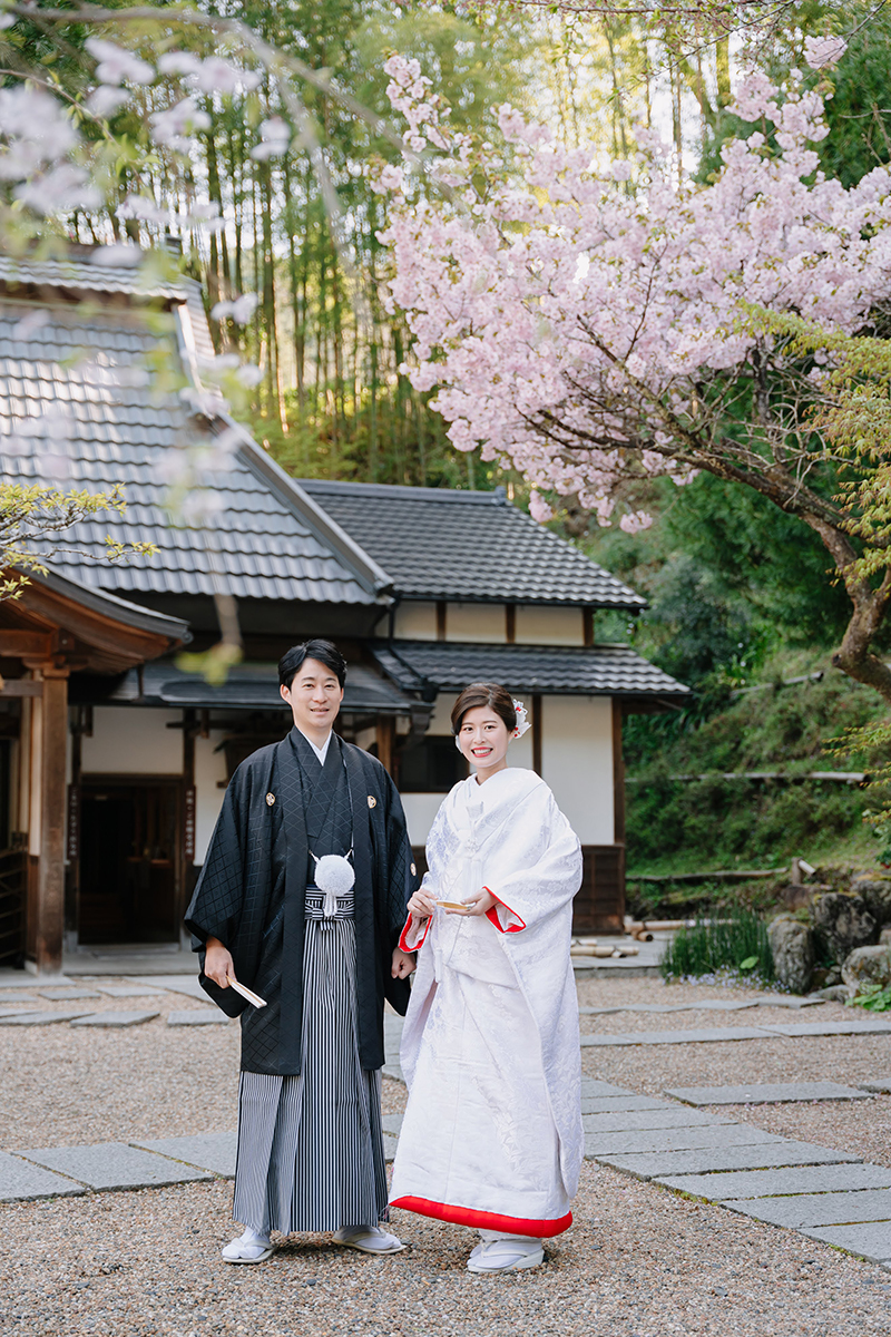 正寿院の境内で赤ふきの白無垢を着た笑顔の新婦と新郎の全身写真