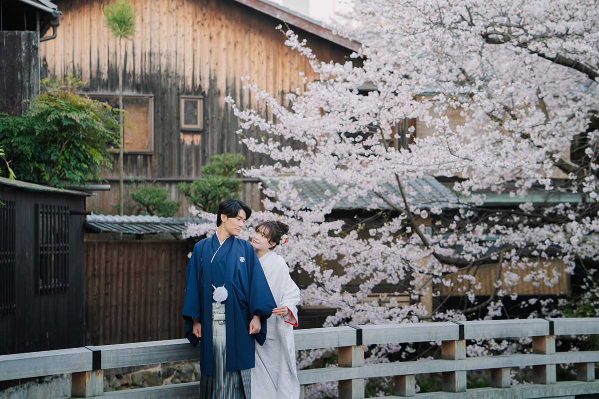 桜バックで赤ふきの白無垢を着ている新婦と紺色の羽織を着ている新郎が楽しそうに話している